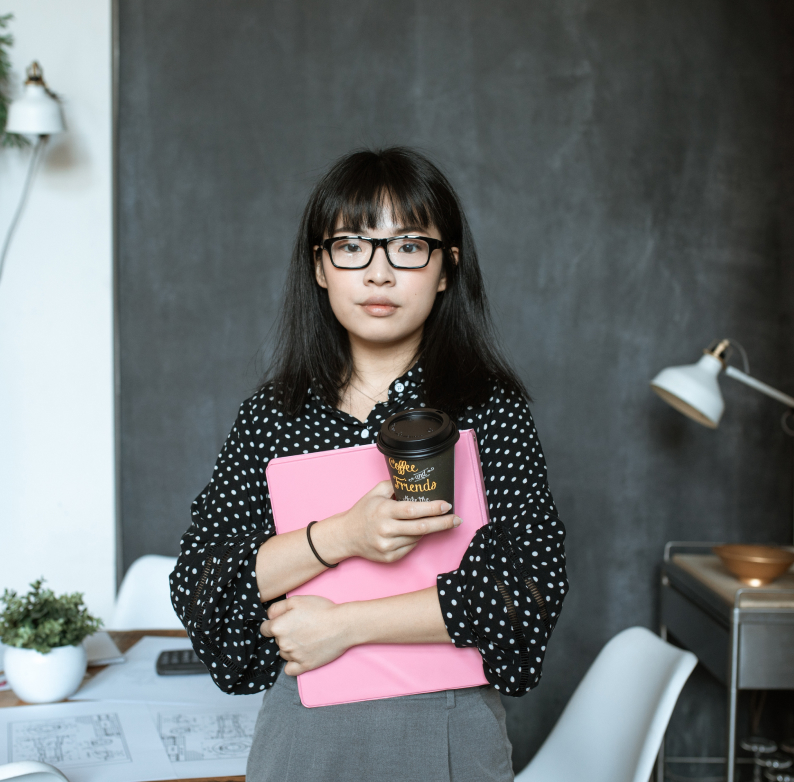 An employee in a polka dotted shirt holding a pink binder and cup of coffee.