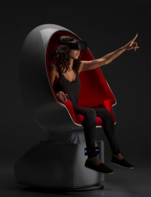 A person sitting in a Positron VR motion chair, wearing a headset, with one finger pointing to the distance.