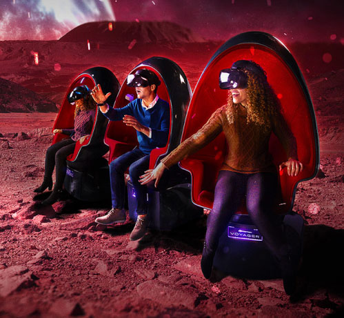 A composite image of three people sitting in Positron Voyager motion chairs. The background is of an reddish alien landscape.