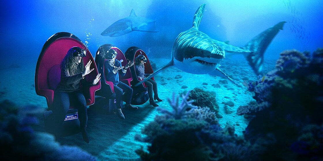 A composite image showing 3 people in Positron motion chairs at the bottom of the ocean, two sharks swimming around them.